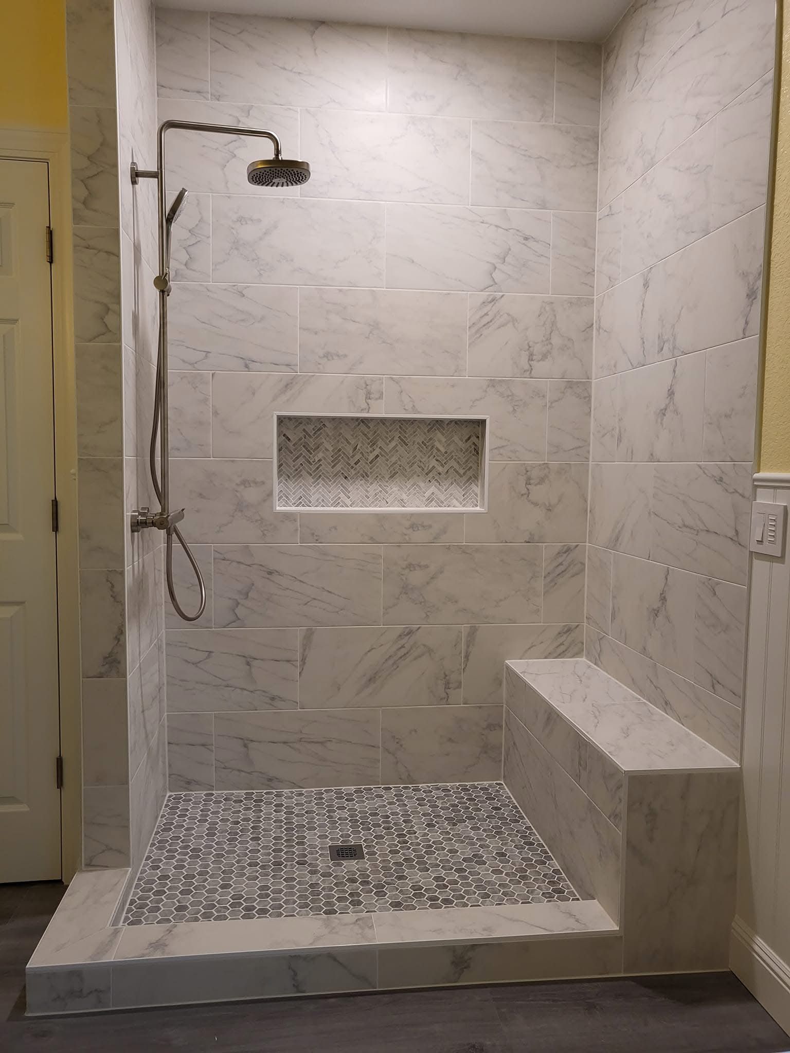 A walk-in shower with marble-look wall tiles, a mosaic tile floor, a built-in bench, and a modern chrome showerhead.