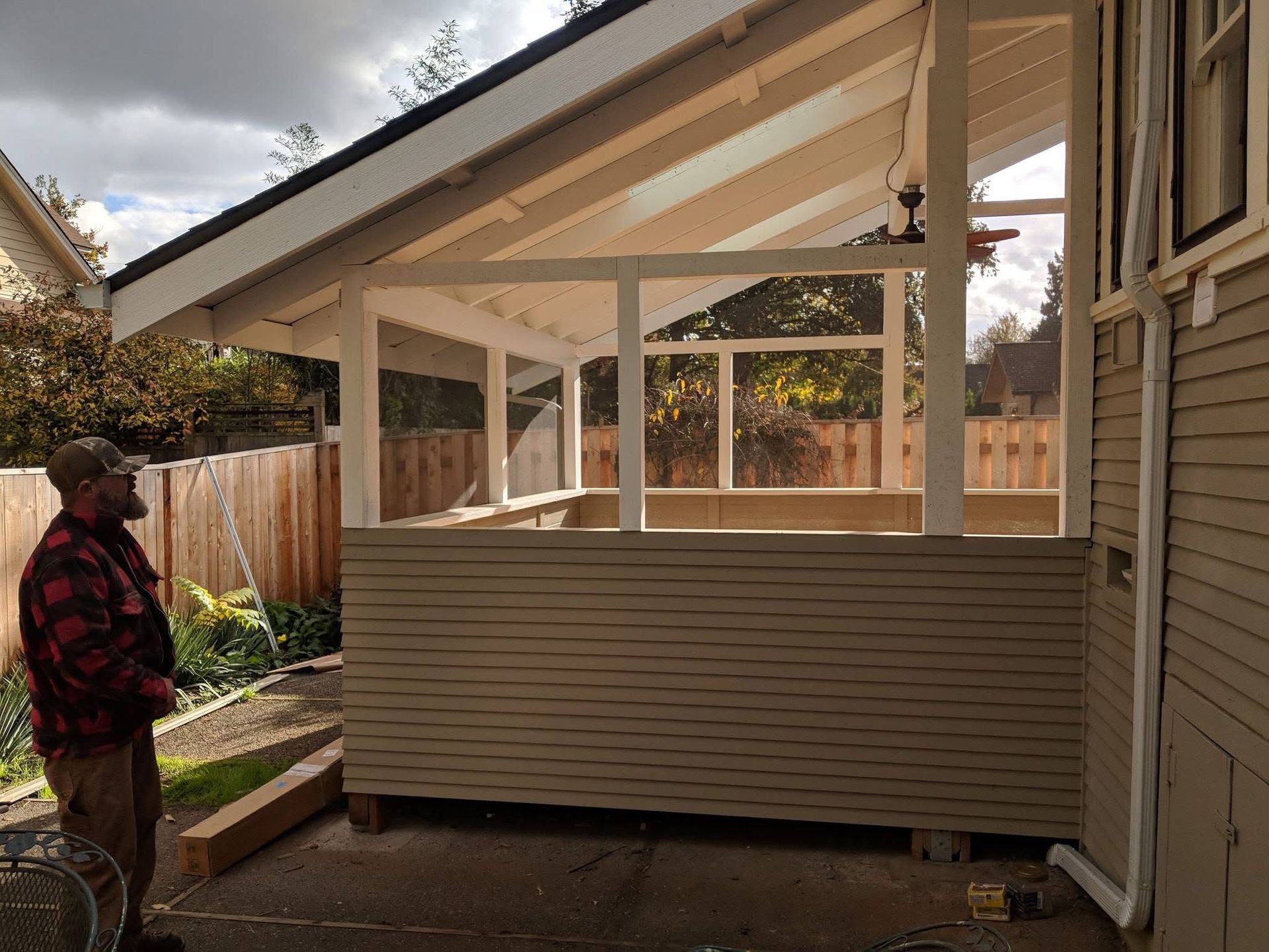 A man in a flannel shirt stands outside next to a partially constructed, light-colored wooden patio enclosure.