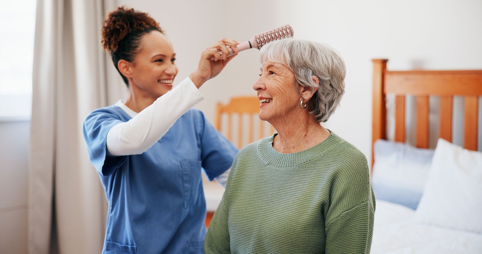 A caregiver in blue scrubs smiles while styling an older individual's hair with a round brush in a bedroom.