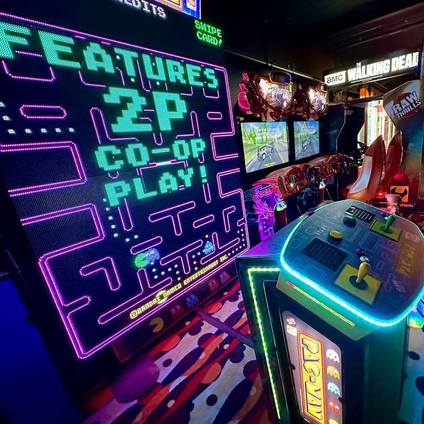 Neon-lit arcade with Pac-Man machine showing “FEATURES 2P CO-OP PLAY” and a side row of arcade cabinets