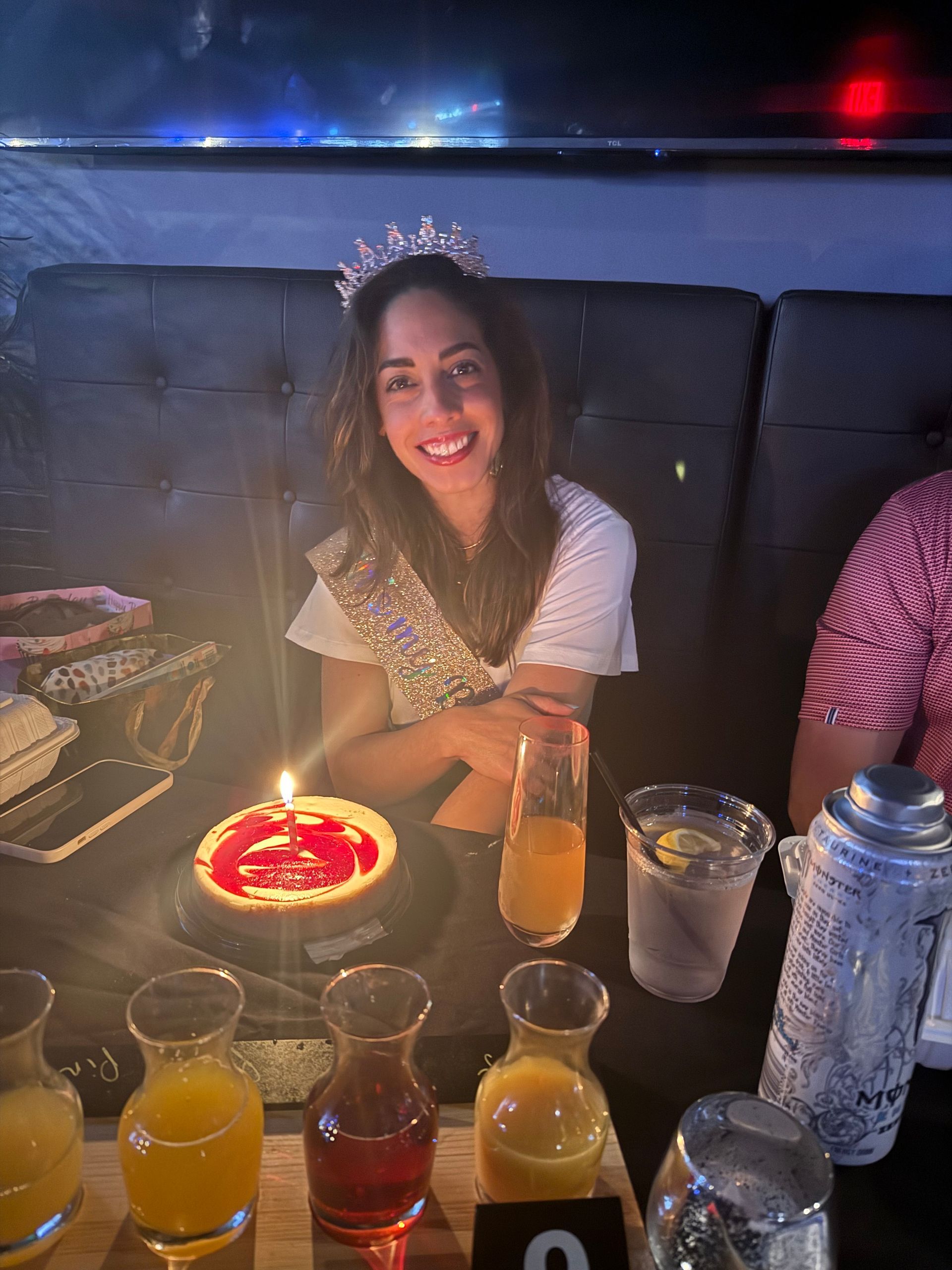 Woman wearing a tiara smiling at a birthday dinner table with a cake and drinks.