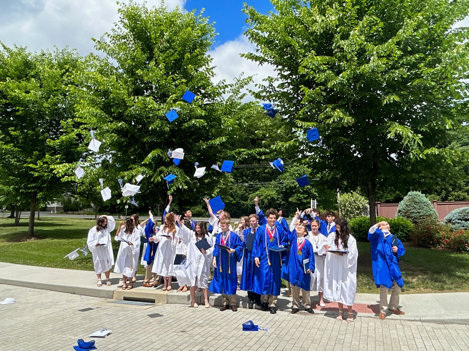 8th grade graduates in blue and white robes toss their mortar board hats high into the air outside 