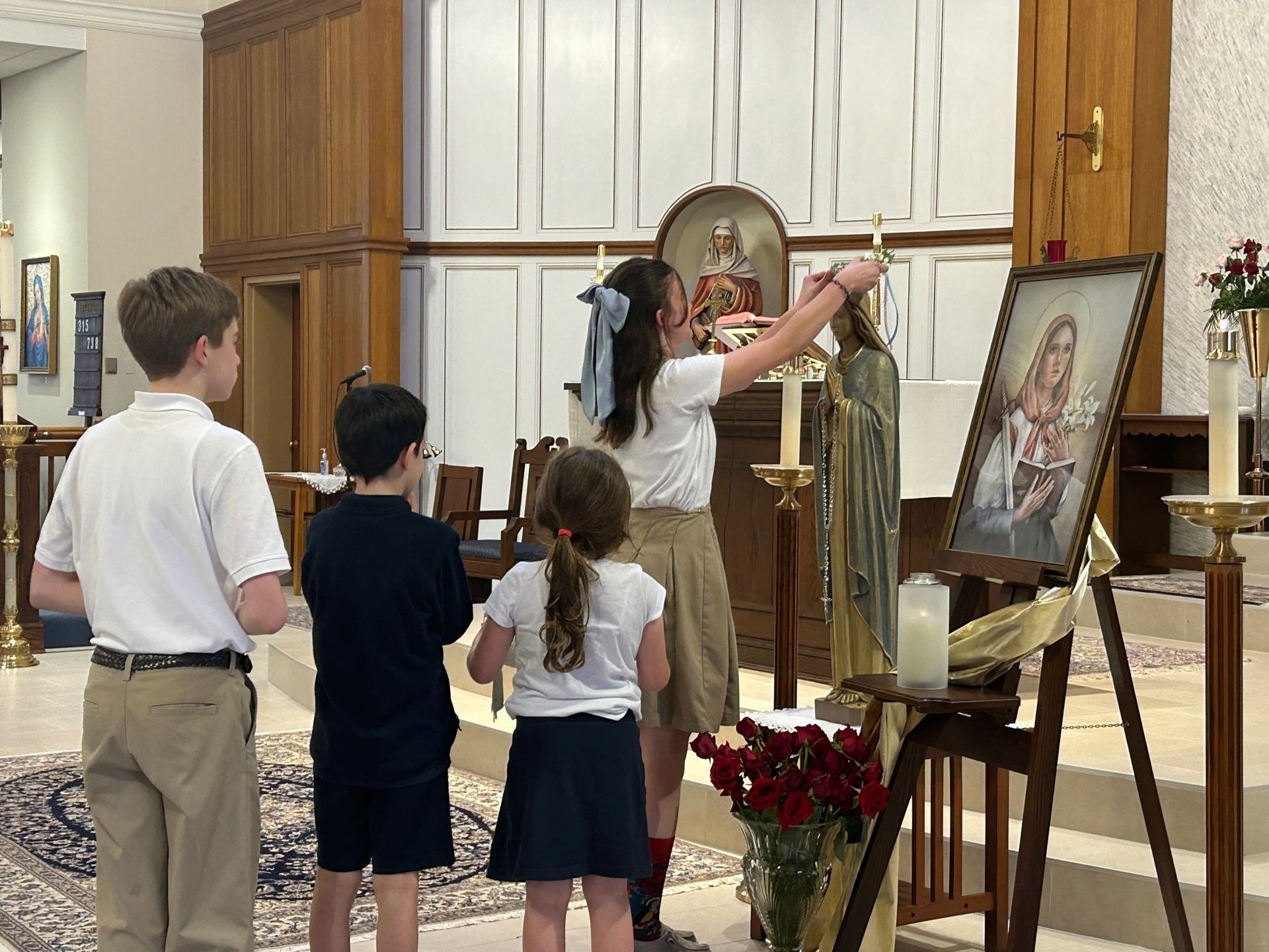 An 8th grade girl places a flower crown on the Mary statue near the altar of the church. Two second grade students and an 8th grade boy stand nearby