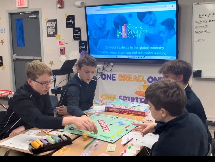 Four middle school boys sit at a table playing Monopoly. A board in the background displays an image that says