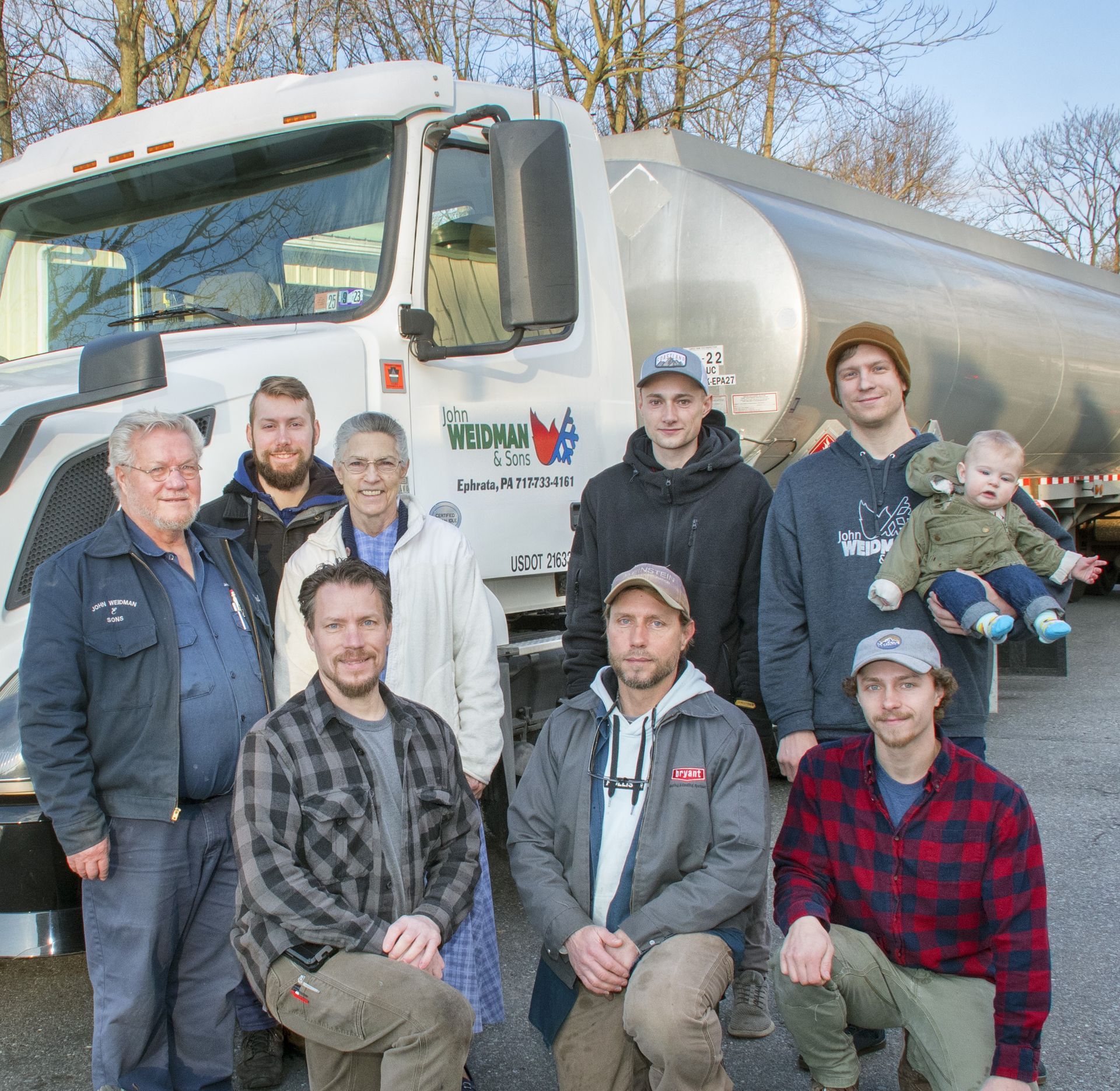 Group of people posing by a white tanker truck. Some wear hats, smiles, and work clothes. A child is held.