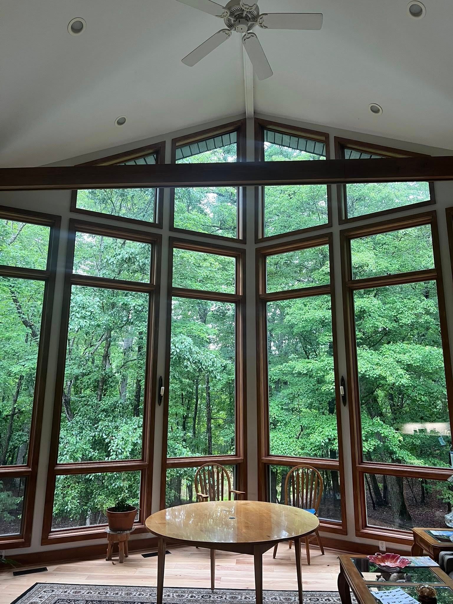 Bright sunroom with floor-to-ceiling windows, wooden chairs, and a round table overlooking lush green trees