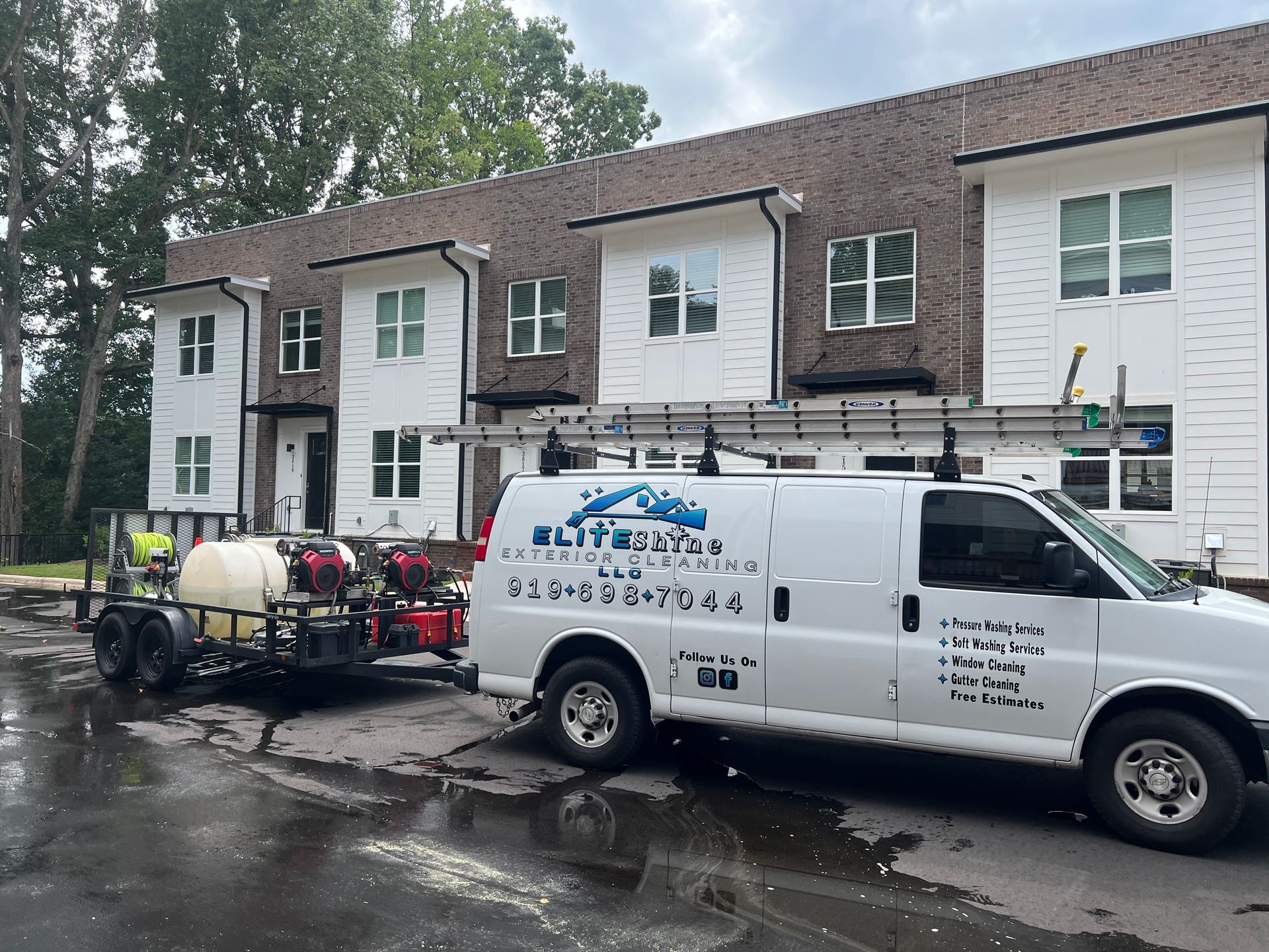 White service van with equipment trailer parked outside a townhouse building on a wet street.