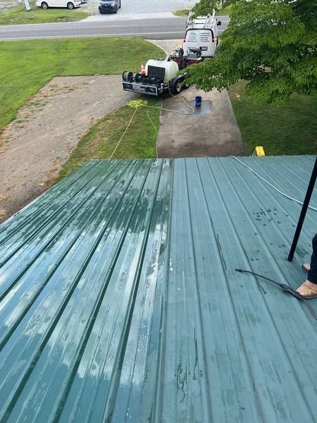 Green metal roof being pressure washed. Half is clean, the other half dirty.  Truck and hose visible.