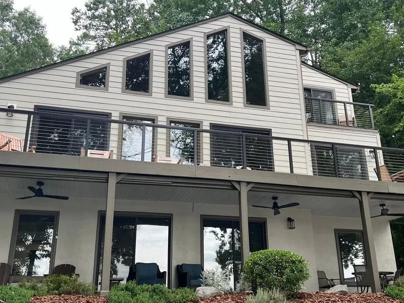 Two-story house with large windows, balconies, and deck overlooking a body of water, surrounded by trees.