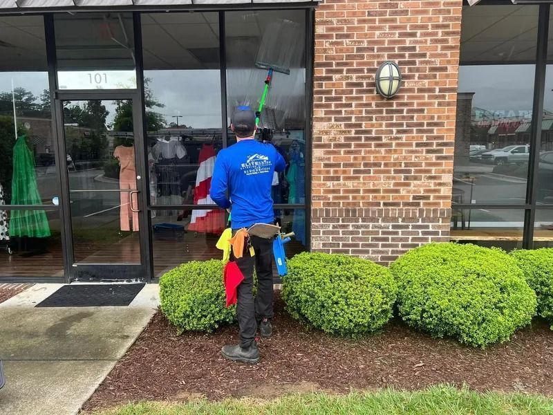 A person cleans windows of a storefront. They wear a blue shirt, stand outside, and use a squeegee.