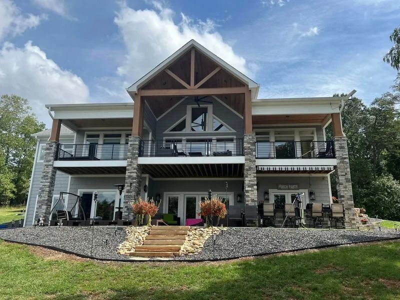 Two-story house with gray siding, stone accents, and balconies, set against a blue sky with green grass and a stone retaining wall.