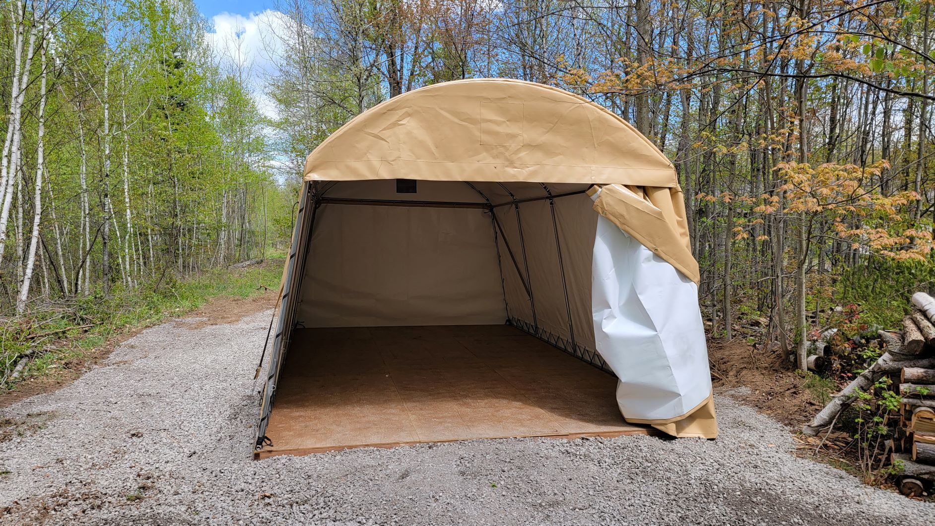 Tan fabric-covered storage shelter on gravel path in a wooded area. The entrance is open.