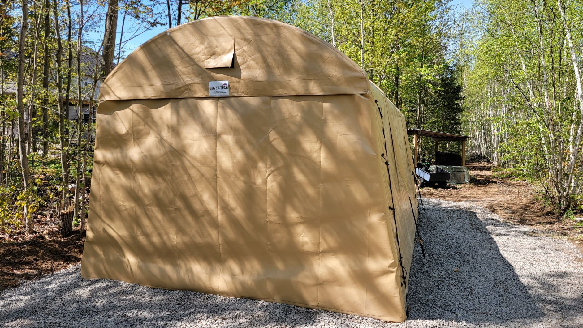 Tan fabric storage shelter on gravel path in a wooded area.