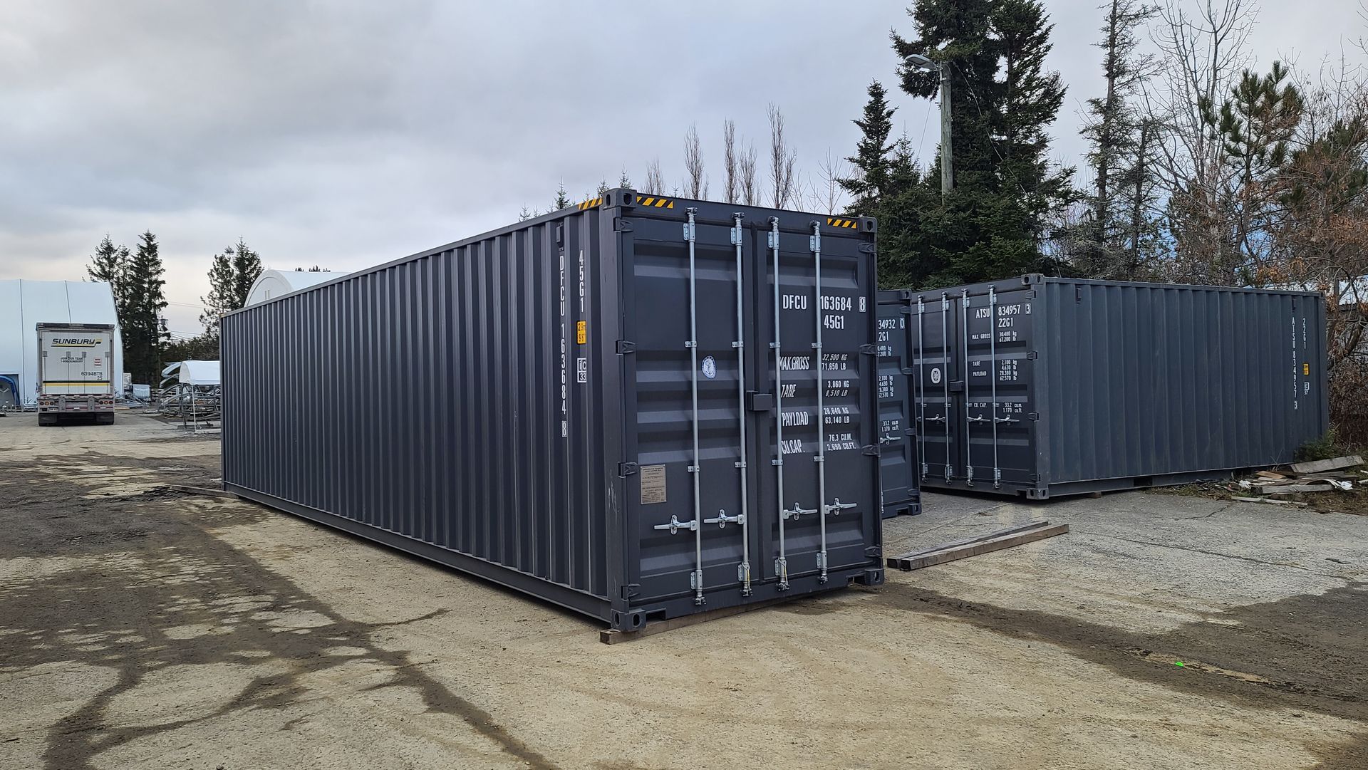 Three dark gray shipping containers on a paved area, with a truck visible in the background. Cover-Tech Shipping Containers.