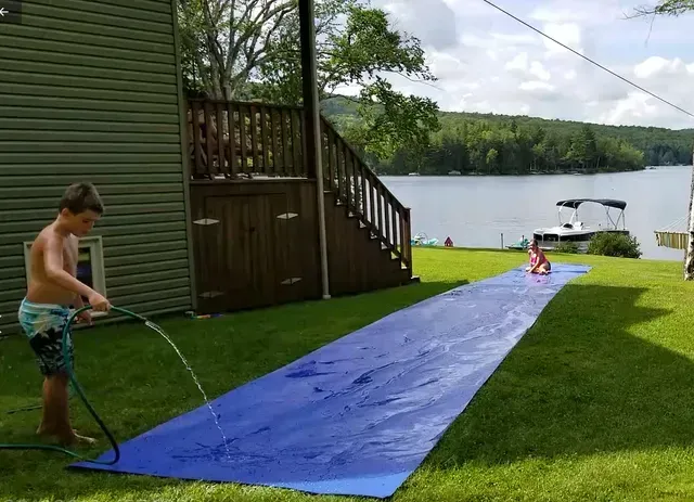 Boy spraying water on a blue slip-n-slide on a grassy lawn; another child slides toward a lake. Cover-Tech Slippy Slope Slip and Slides,