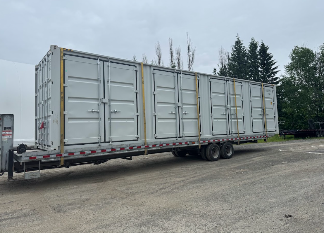Cover-Tech White shipping containers on a flatbed trailer, secured with straps. Outdoors, overcast day.
