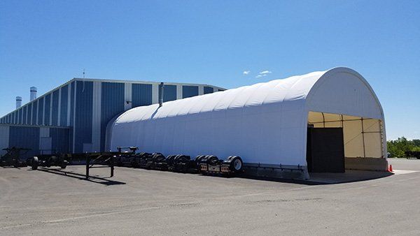 White arched-roof storage structure in front of a blue and white industrial building on a sunny day. Cover-Tech Replacement Covers.