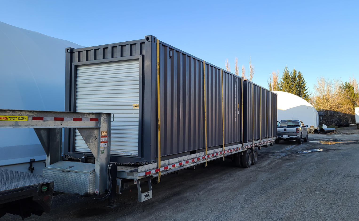 A large, dark gray Cover-Tech shipping container with a roll-up door is secured to a flatbed trailer outdoors.