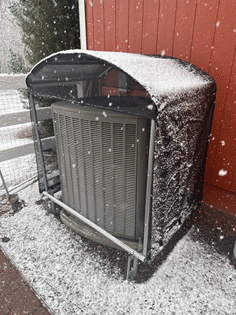 AC unit covered with snow, under a protective canopy, next to a red building during a snowfall.
