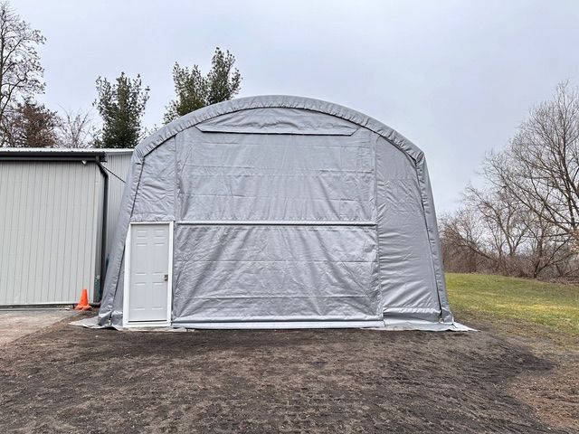 Gray arched-roof storage structure with a roll-up door, a white side door, and a white building alongside.