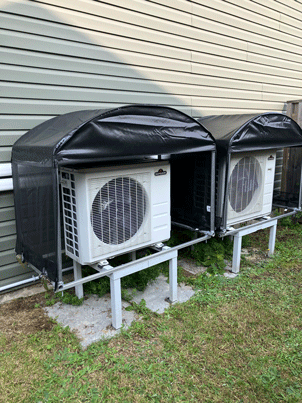 Two air conditioning units with black covers, mounted on metal stands, near a house.