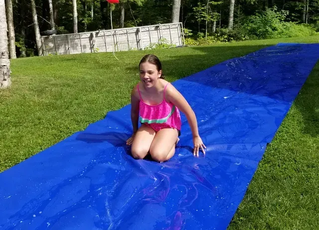 Girl kneels on a blue slip-n-slide on green grass. She wears a pink swimsuit, smiling. Water on the slide. Cover-Tech Slippy Slope Slip and Slides,