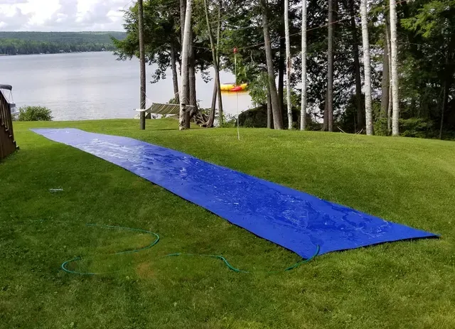 Blue slip-n-slide on green lawn, overlooking a lake with trees. Cover-Tech Slippy Slope Slip and Slides,