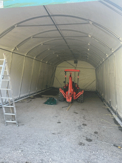 Inside a fabric-covered structure: a small orange tractor is parked on gravel. A ladder leans to the left.