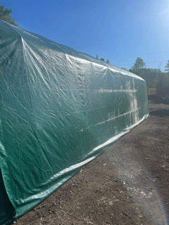 Green tarp-covered structure outdoors against a blue sky, possibly a greenhouse or storage shed.