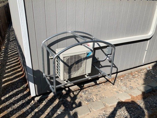 Metal cage protecting an air conditioning unit mounted on a gray shed wall, gravel ground.
