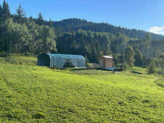 Green field with a large, arched structure and a small orange shed in front of a forest and hills.
