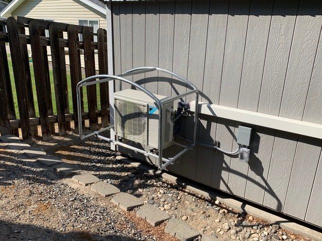 Air conditioner unit with protective metal cage mounted on a light gray building wall, gravel ground, and wooden fence.
