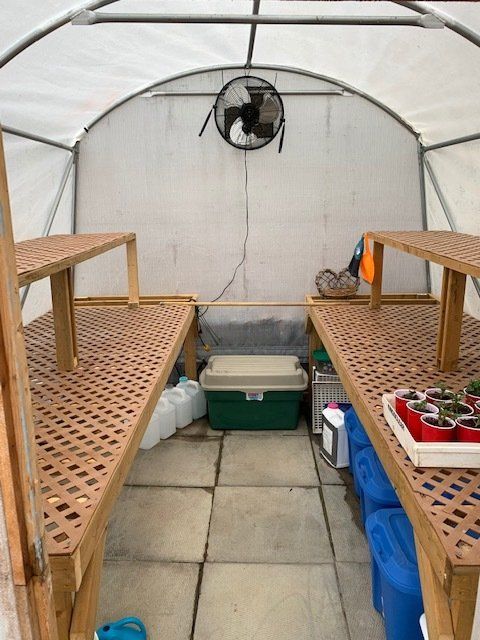 Interior of a greenhouse with two wooden workbenches, a fan, and storage containers.