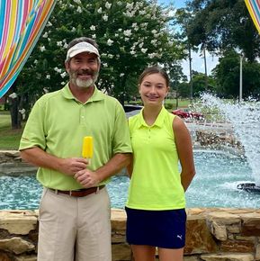 Man and young person in lime green shirts pose near a fountain. Man holds a popsicle.