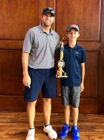Man and boy holding trophy, smiling. Man in striped shirt and shorts, boy in blue shirt and gray shorts. Wooden background.