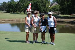 Four golfers standing on a green with golf clubs, posing by a pond, under a waving flag.