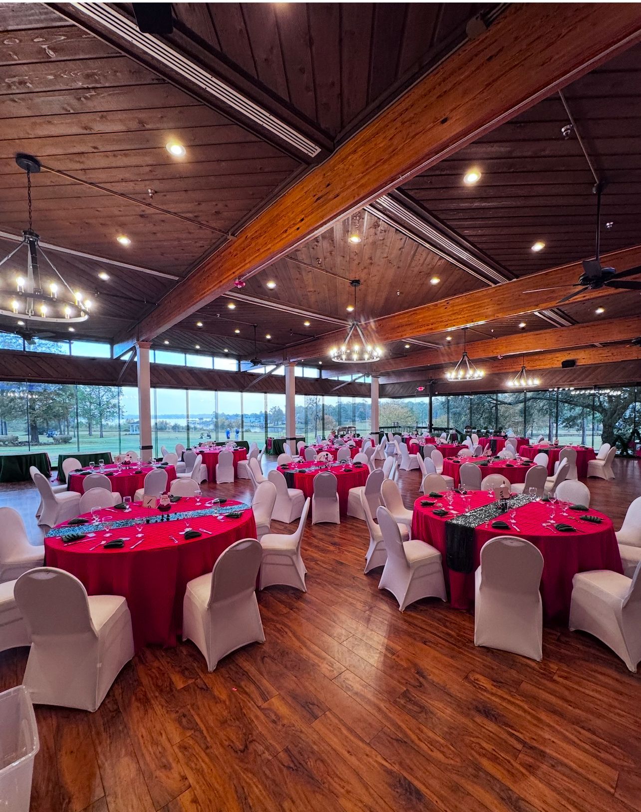 Reception hall with round tables covered in red linens, white chairs, wooden floor, large windows.