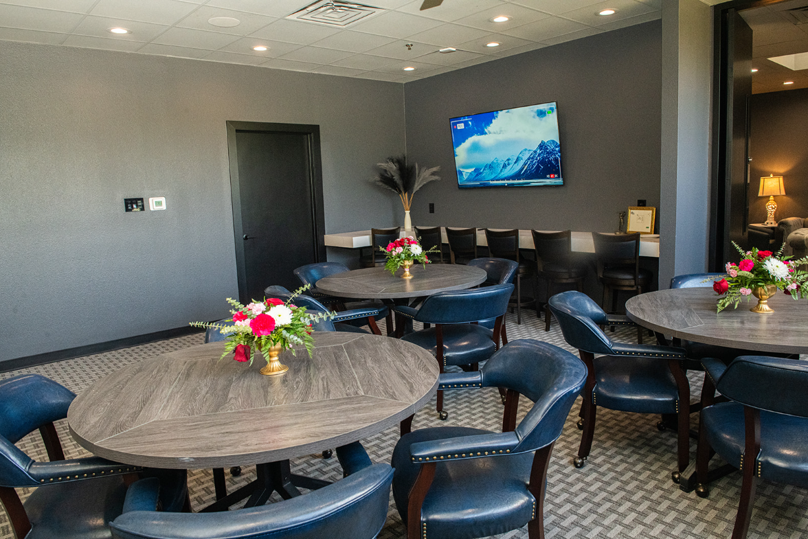 A modern lounge with round tables, blue chairs, flowers, and a large TV. Dark gray walls and a window.