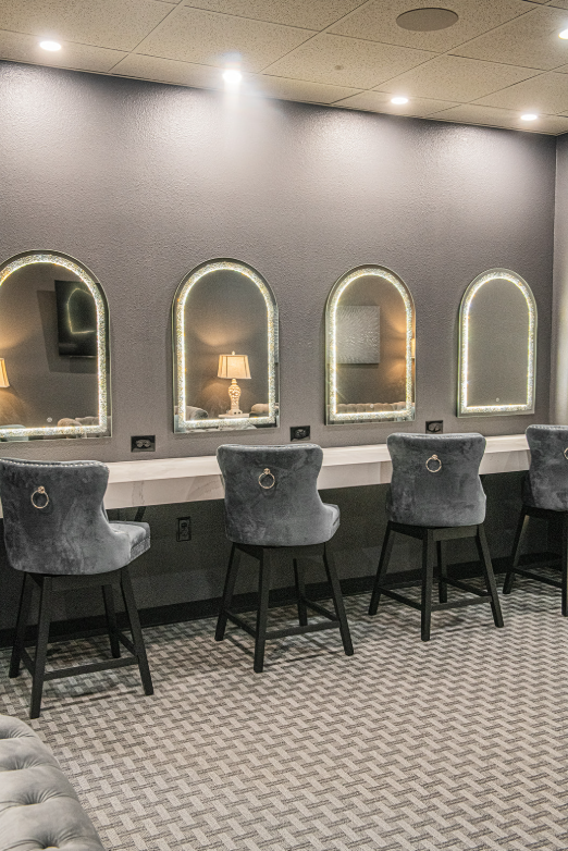 Vanity room with gray walls, arched mirrors, lighted frames, stools, and a long counter.