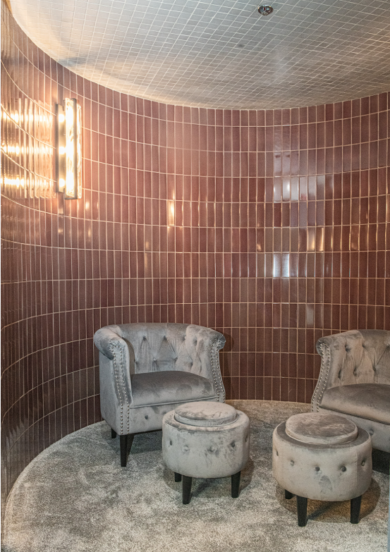 Curved room with reddish-brown tiled walls, gray velvet chairs and ottomans on a gray carpet.