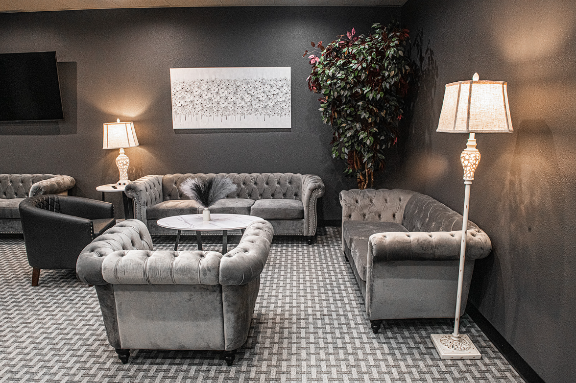 Cozy lounge area with gray tufted sofas, armchairs, lamps, and a decorative tree against a dark wall.