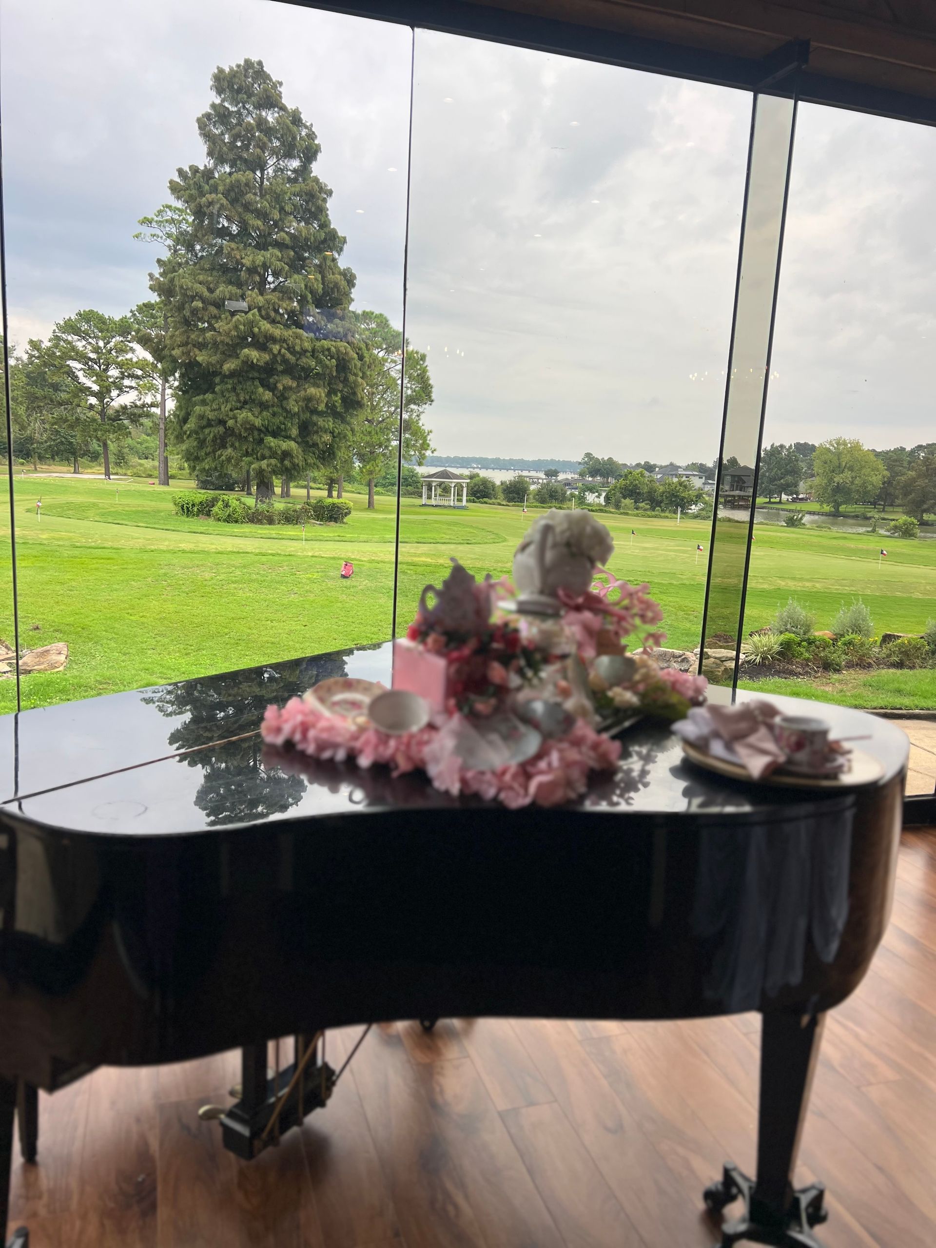 Black grand piano with floral arrangement, large window with outdoor view of green field and trees.