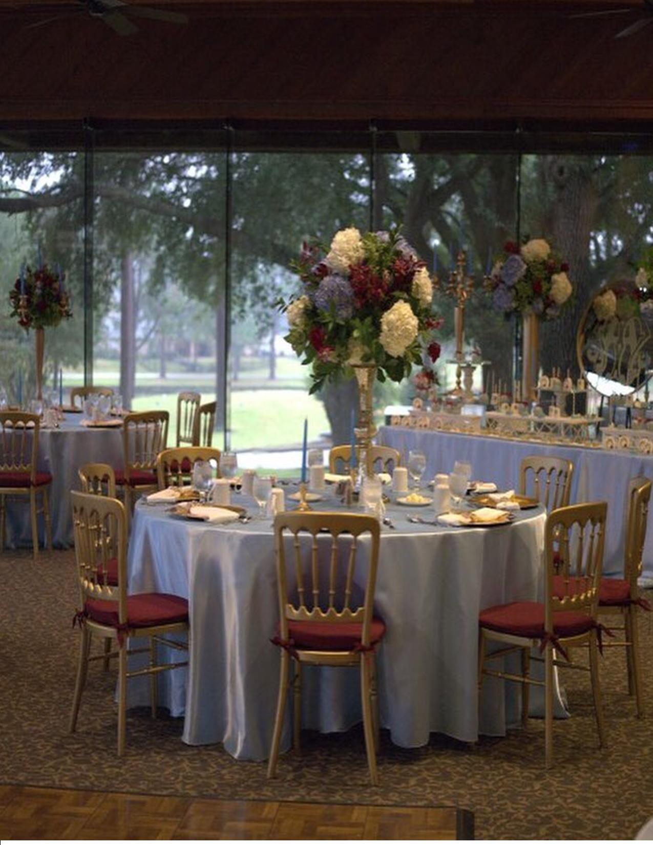 Round tables set for a formal event, with floral centerpieces and a large window overlooking trees.