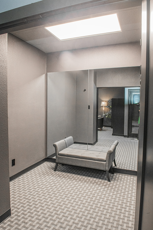 Hallway with a bench, looking into another room. Gray walls, light fixture, patterned carpet.