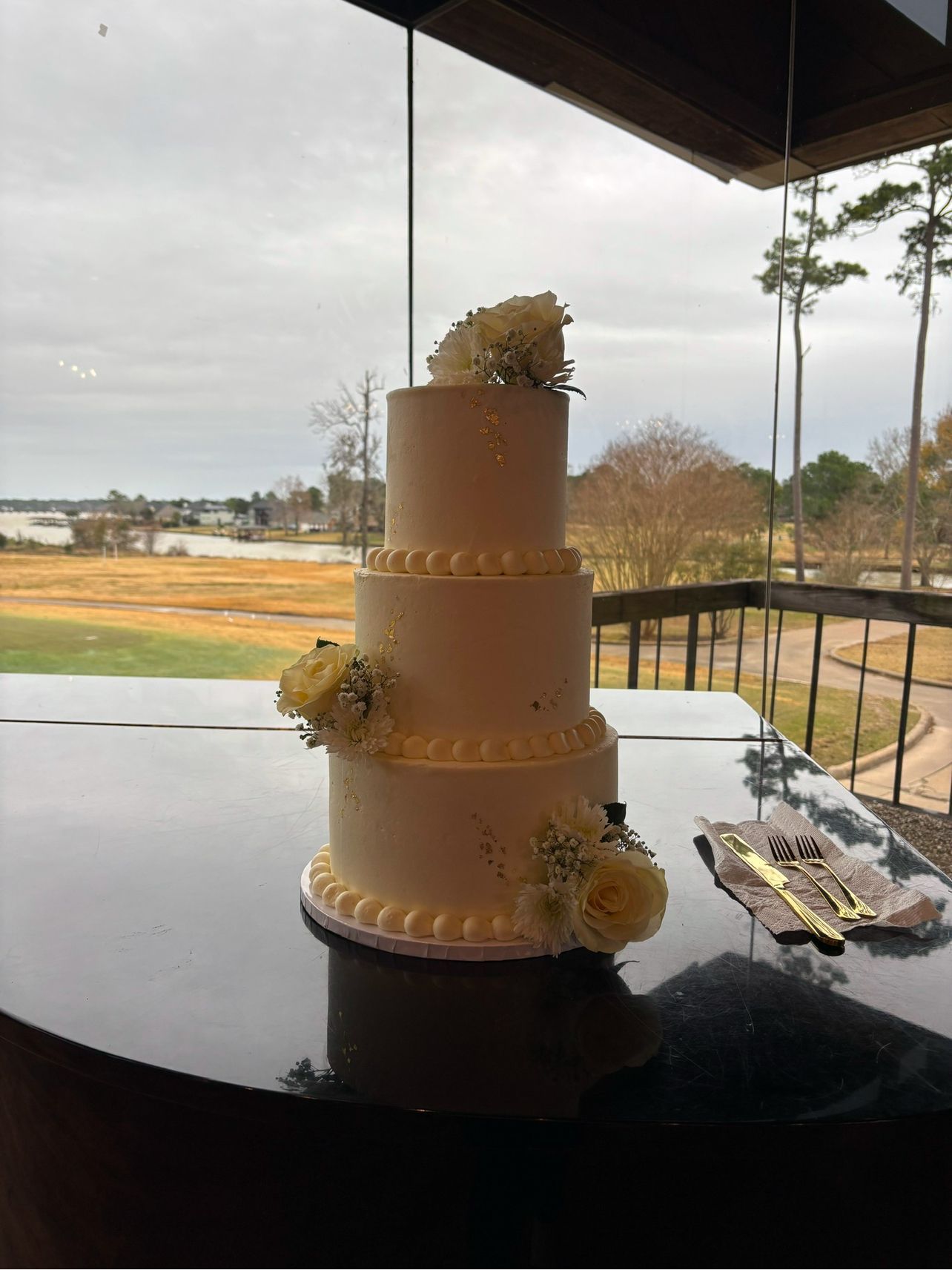Three-tiered wedding cake with white frosting and flowers, on a table by a window overlooking a landscape.