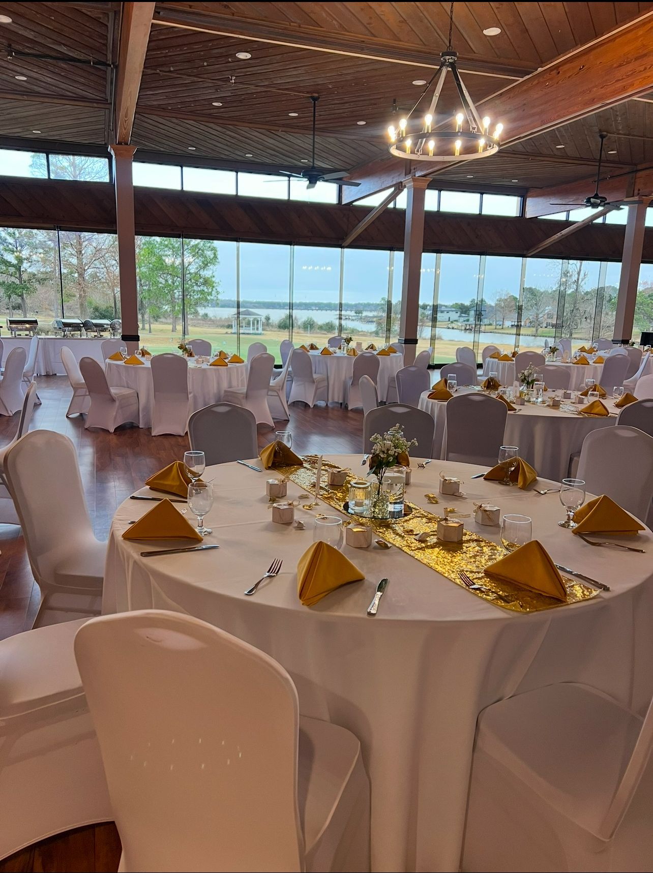 Wedding reception setup in a venue with large windows. Round tables with white linens, gold accents, and white chair covers.