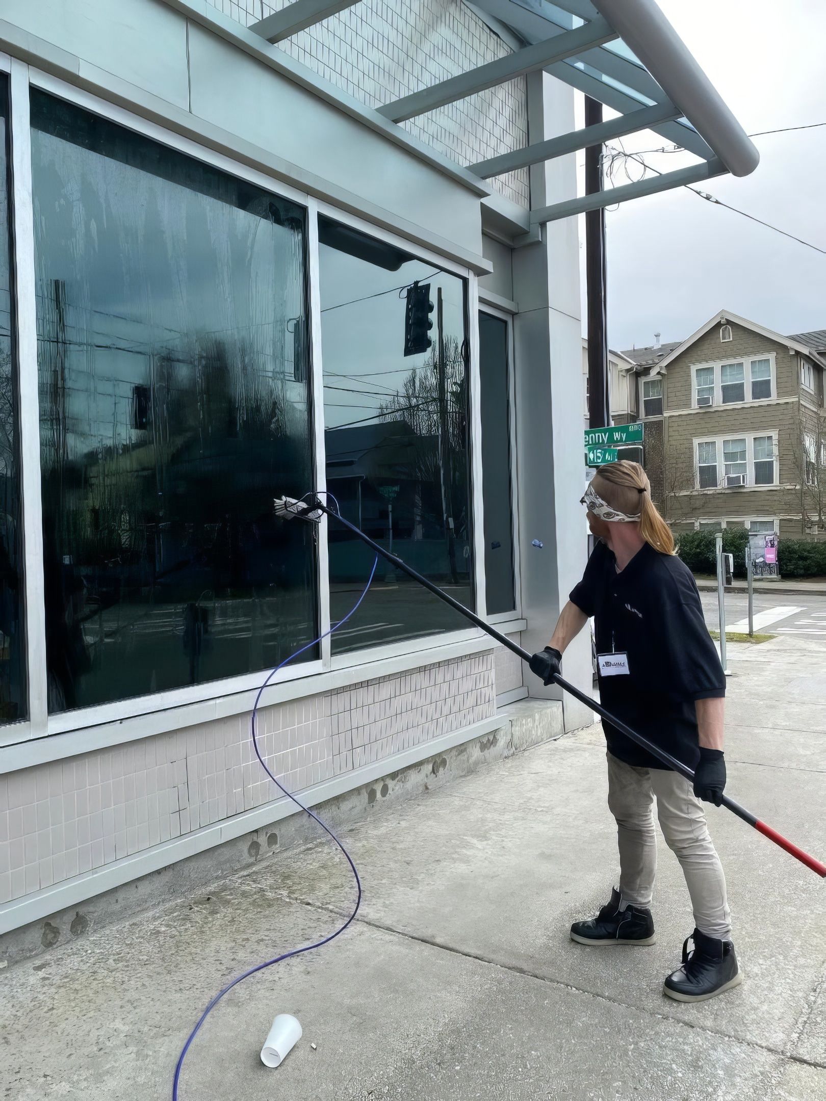 Person pressure-washes the exterior wall of a building beside large windows.