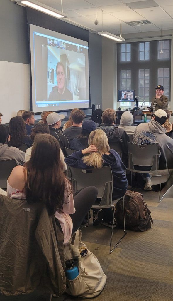A group of students in a lecture hall watch a video on a projector screen. A person stands nearby.