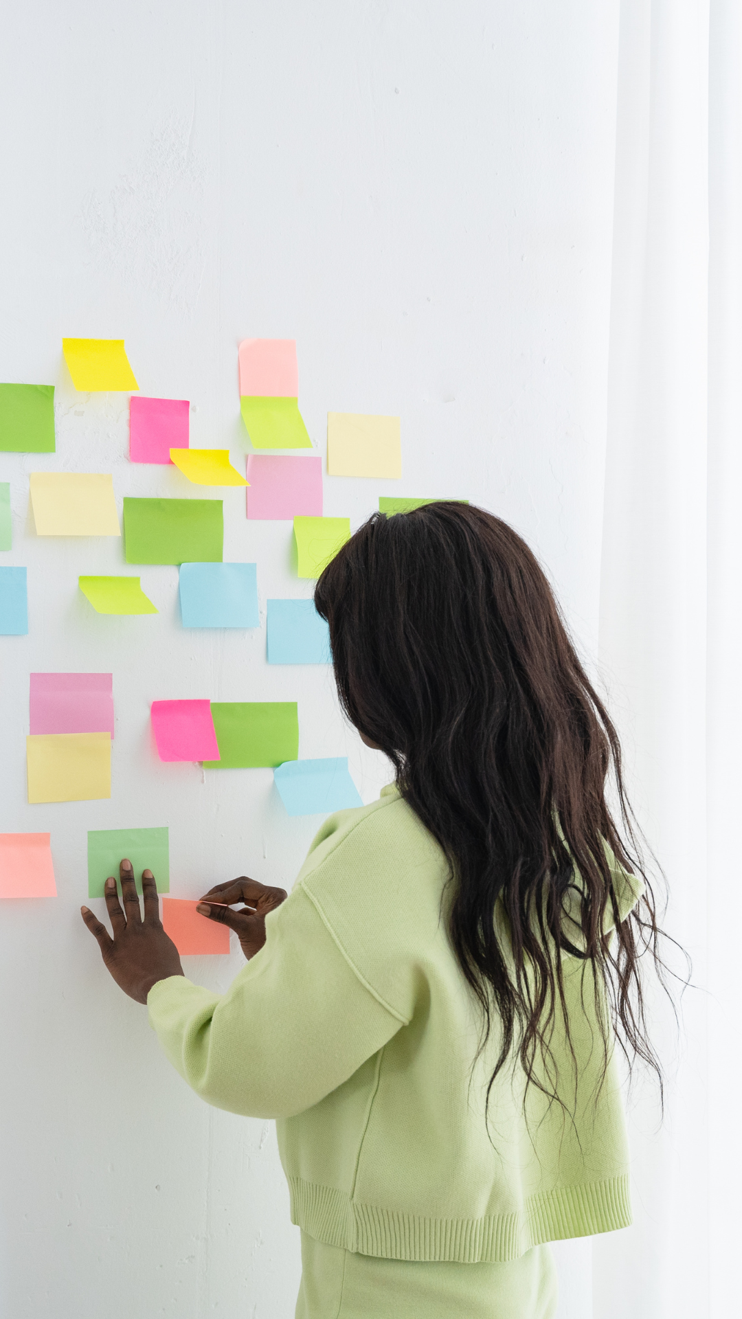 Woman with dark hair in a green outfit, arranging colorful sticky notes on a white wall.