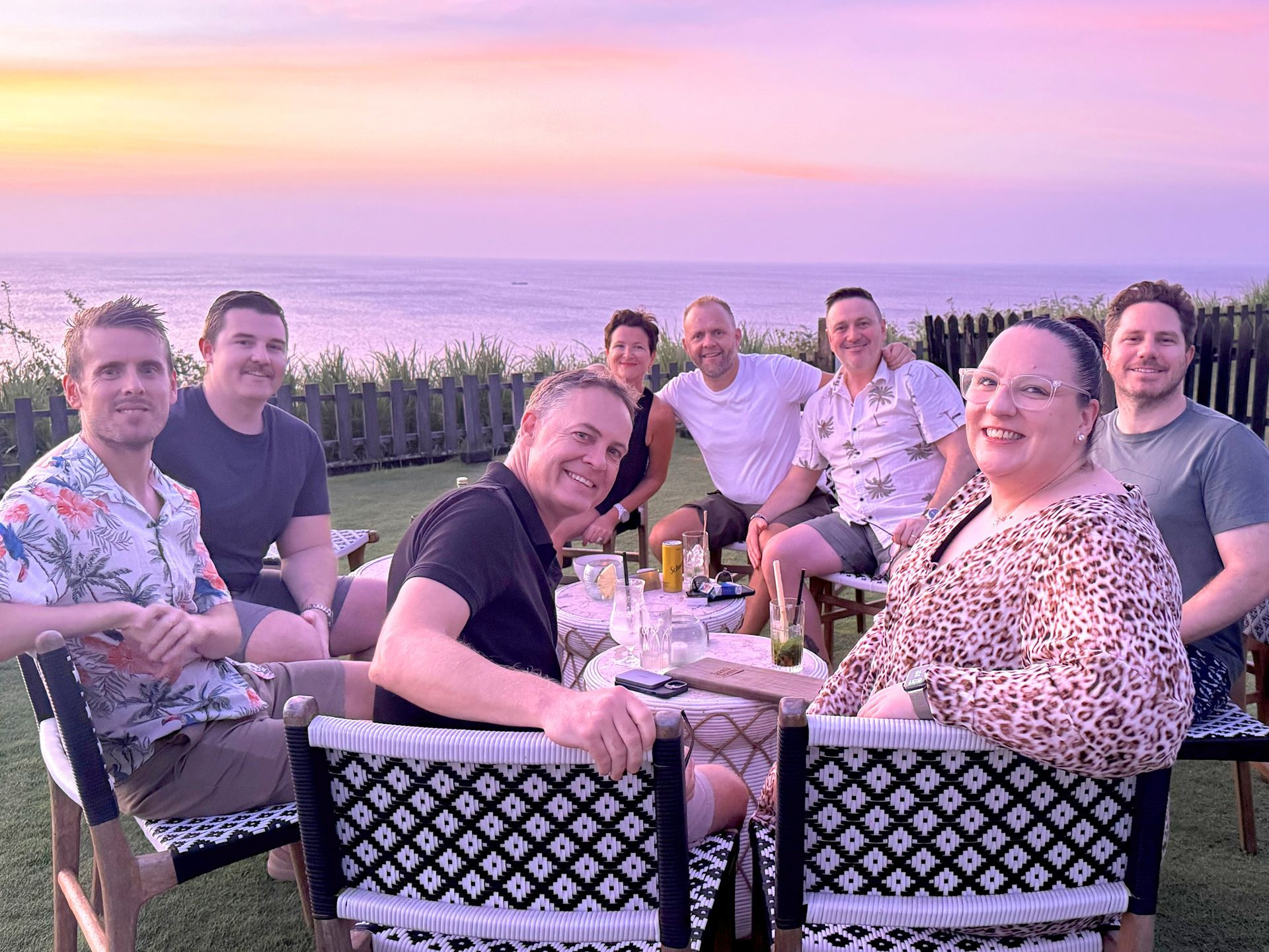 Group of colleagues smiling at an outdoor table by the water at sunset, with pink sky and drinks.